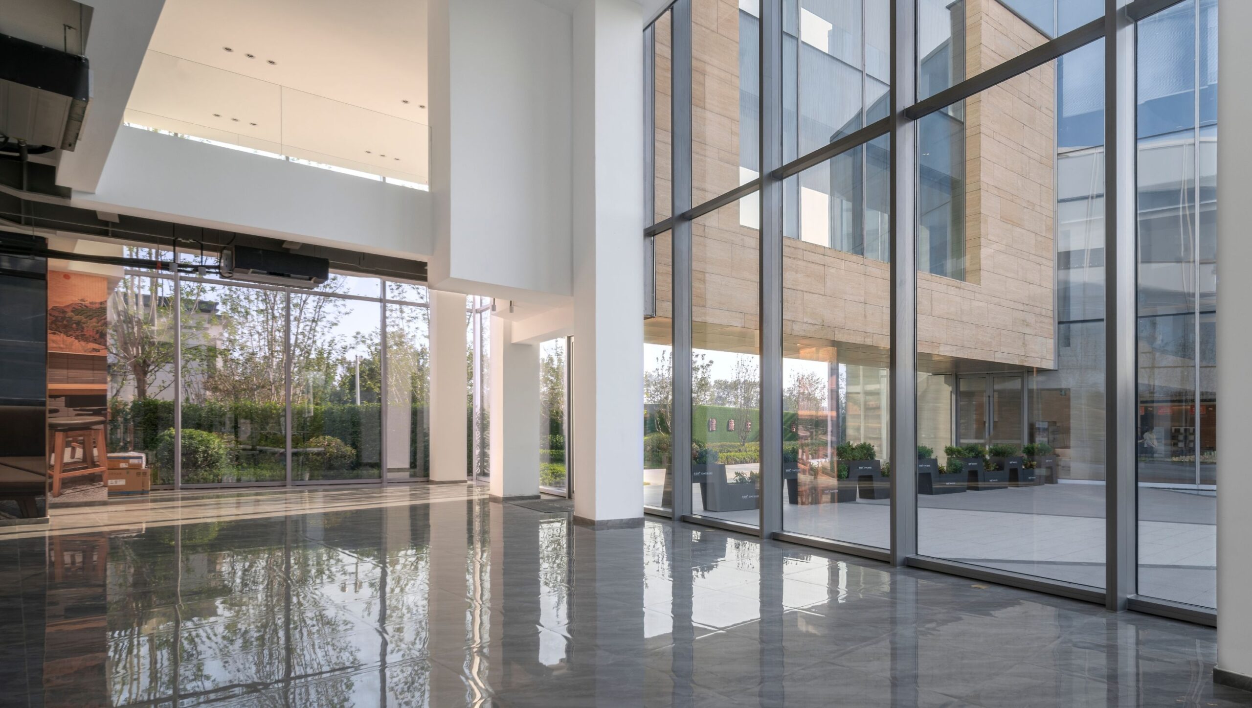 Modernes, lichtdurchflutetes Foyer mit großen bodentiefen Glasfenstern, glänzendem Steinboden und Blick auf einen begrünten Innenhof mit modernen Gebäudefassaden.